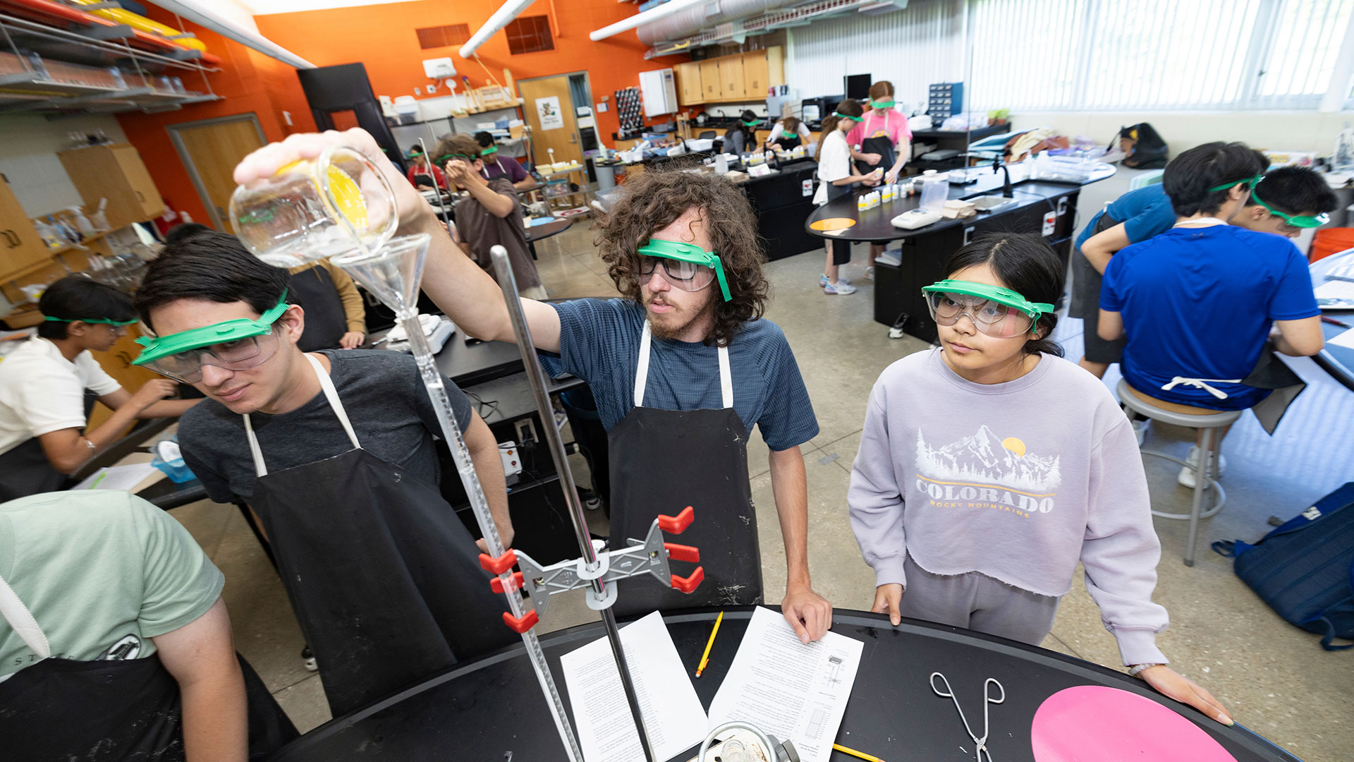A high schooler at Greenwood Laboratory School pours a liquid into a funnel during chemistry class as his peers watch at his side.